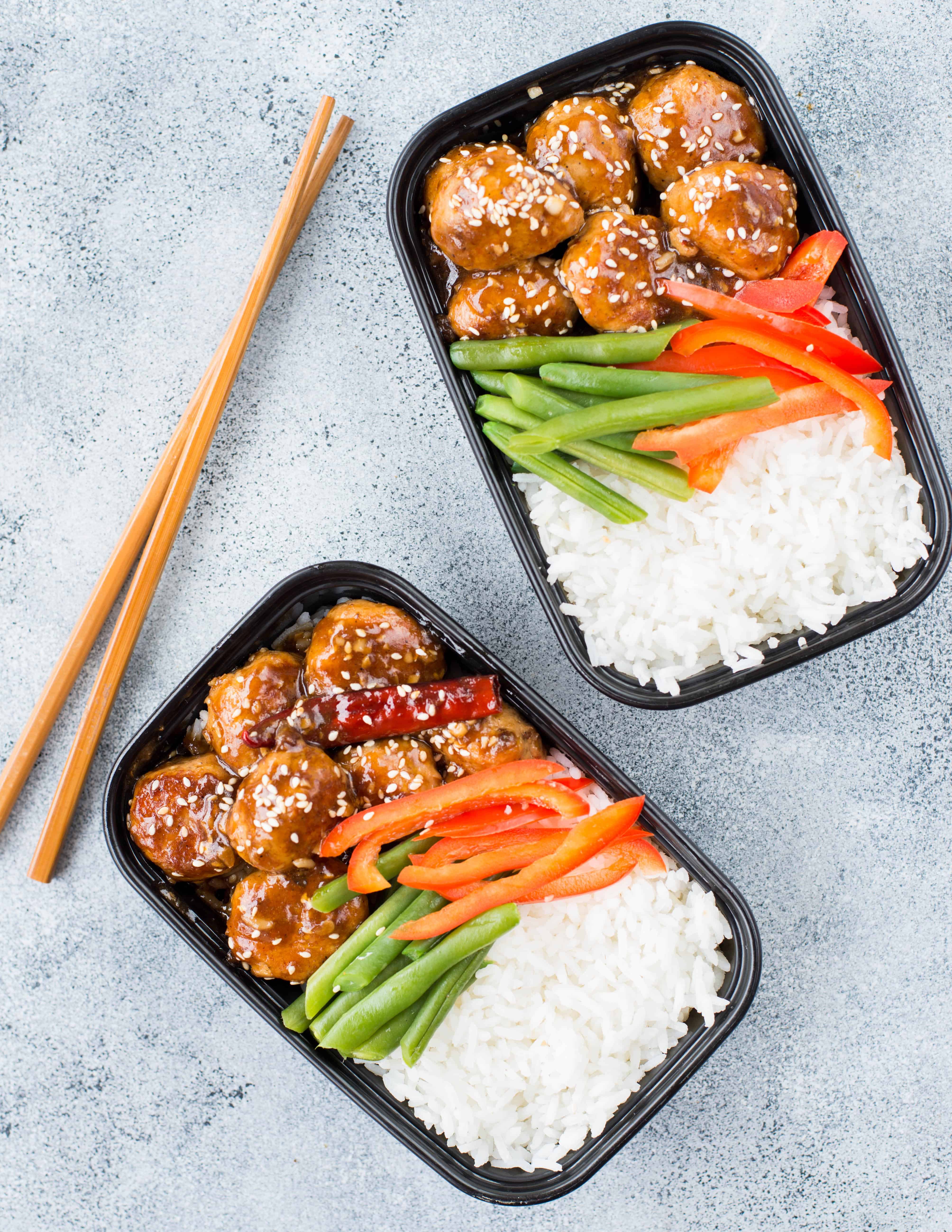 Couple of meal prep lunch boxes containing chicken meatballs with asian sauce, white rice and some greens like beans and red peppers.