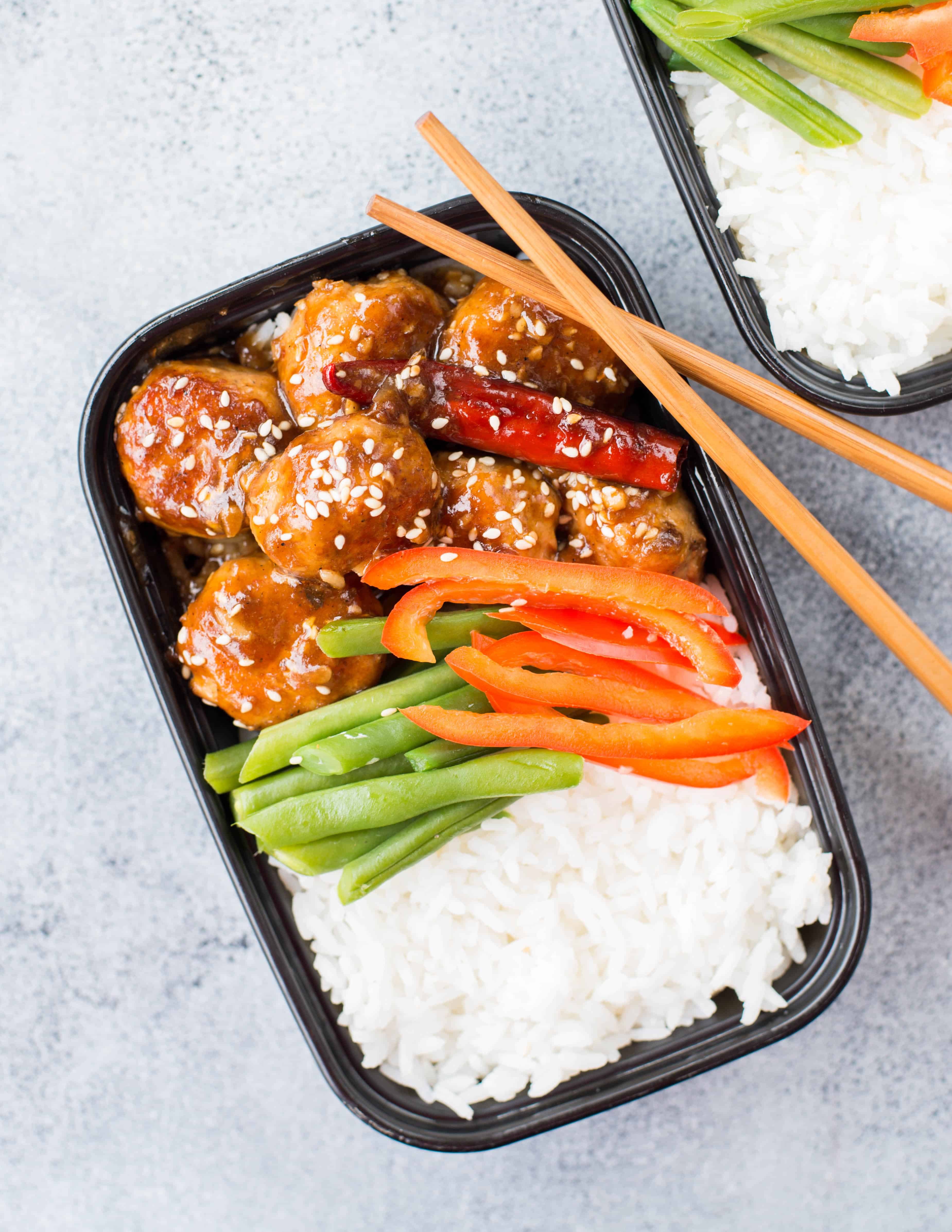 Close up photo of Meal prep lunch box containing chicken meatballs with asian sauce, white rice and some greens like beans and red peppers.