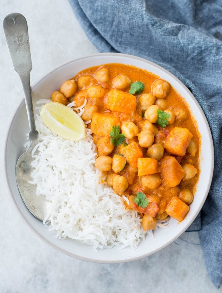 Sweet potato chickpea curry served in a white bowl with a spoon and has fresh cilantro and lemon slice as garnish.