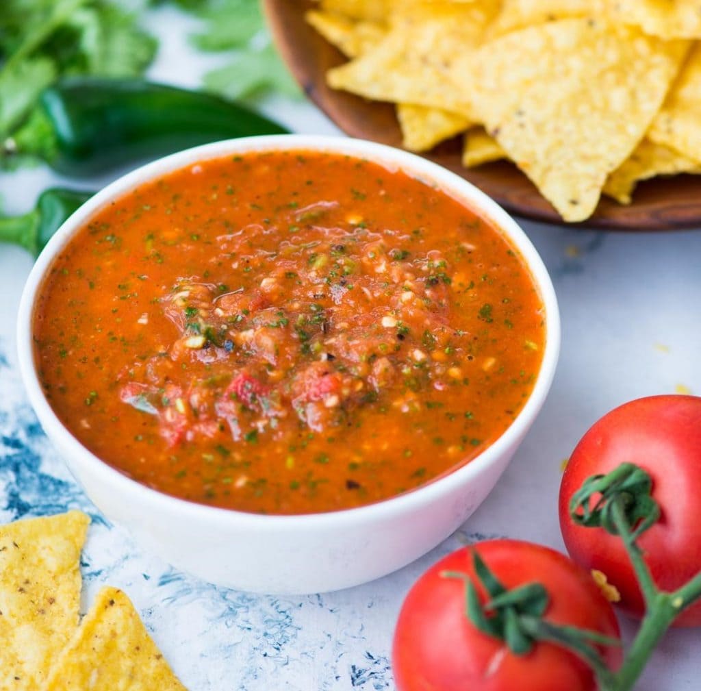 Side view of tomato salsa shown in a white cup alongside tortilla nachos and highlights tomatoes as primary ingredients.