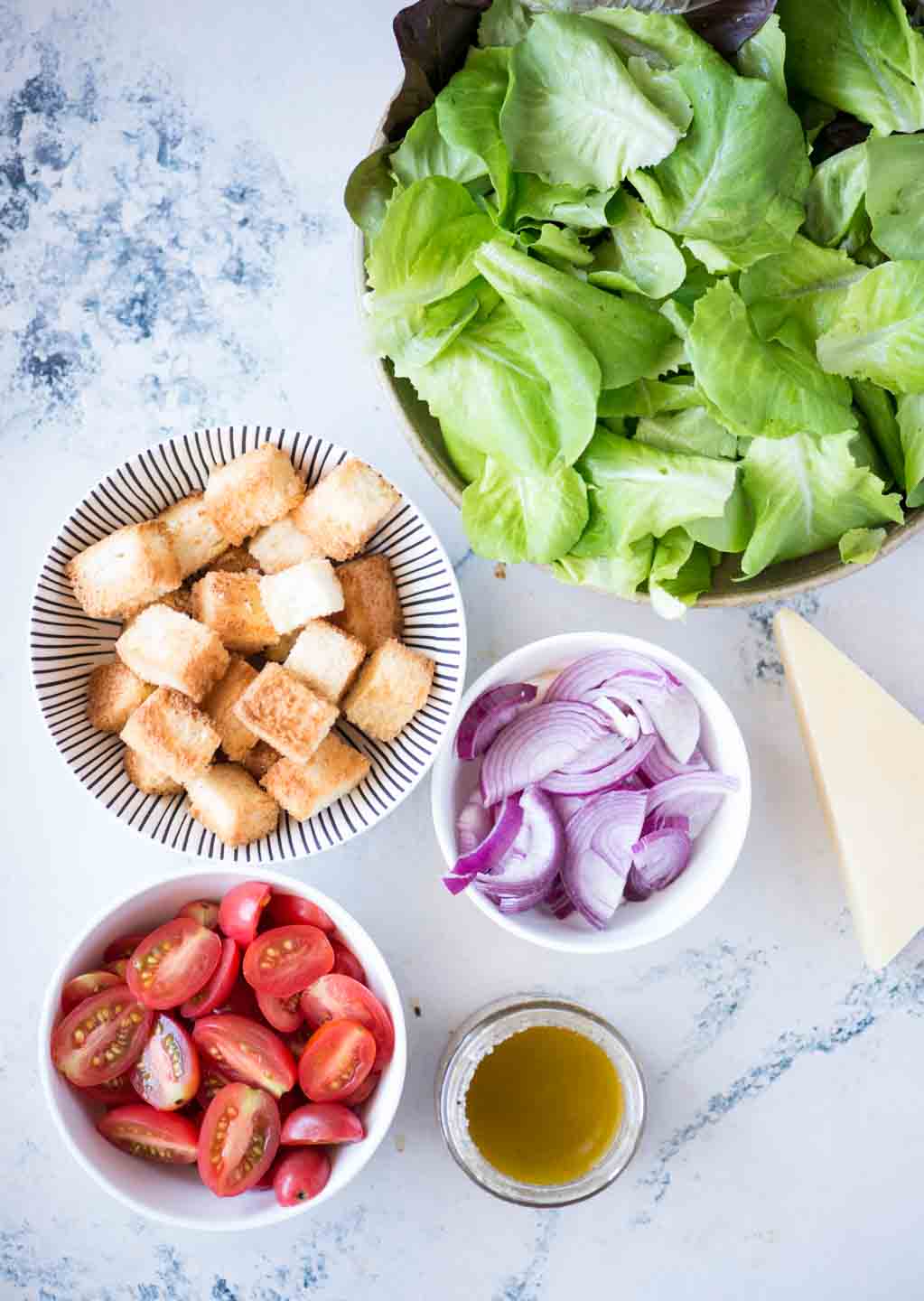 Chopped lettuce, cherr tomatoes, sliced onion, croutons, olive oil and parmesan cheese block shown as ingredients required to make Italian chopped salad.
