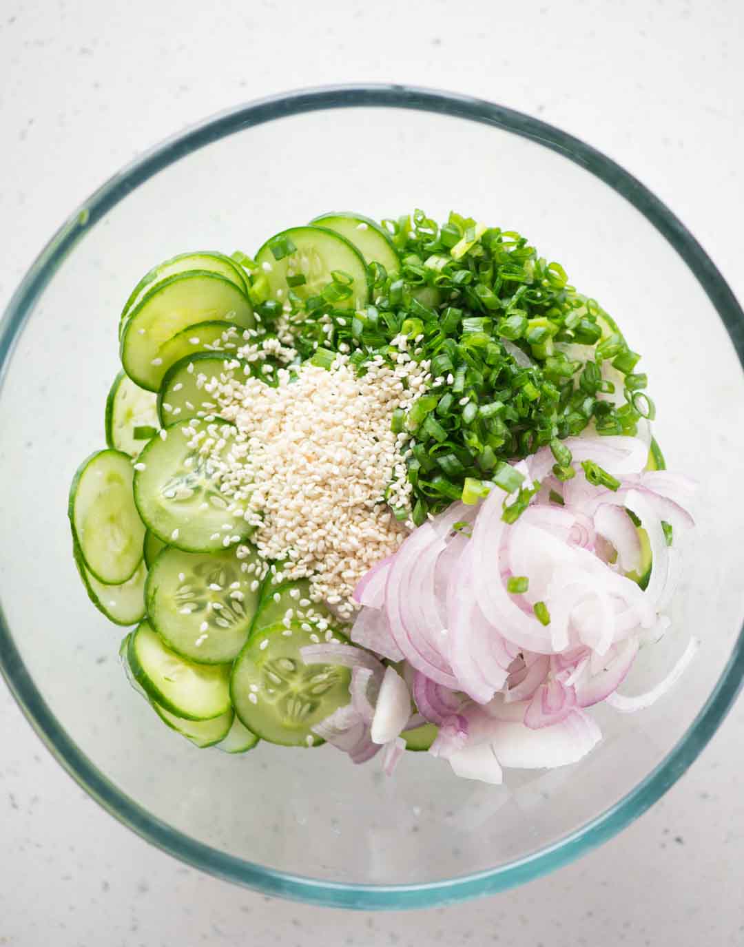 Sliced cucumber and onions with chopped green onions and toasted sesame seeds in a large bowl to make cucumber salad.