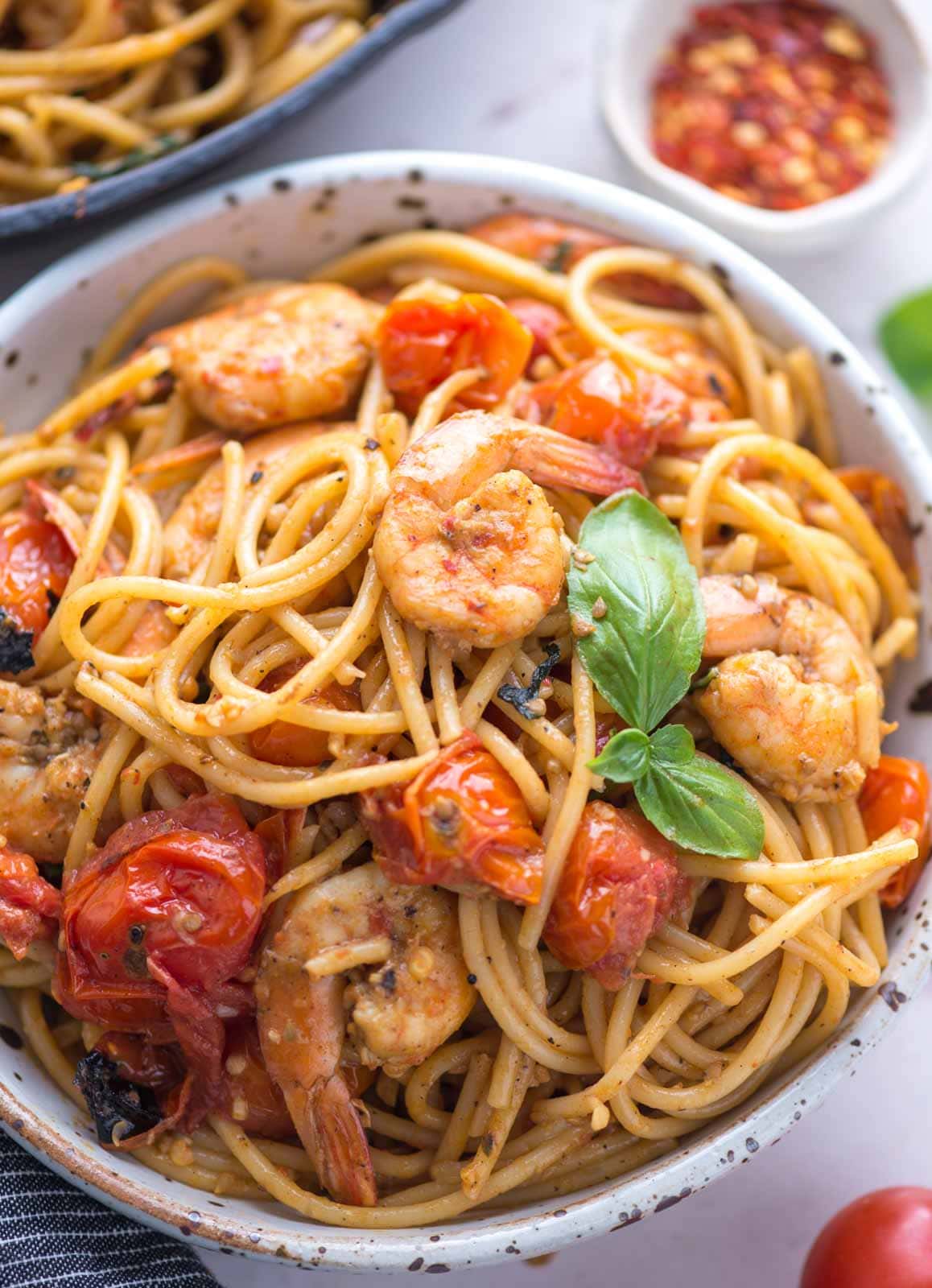 A bowl of pasta showing fried shrimps, cooked crushed cherry tomatoes and fresh basil on top.