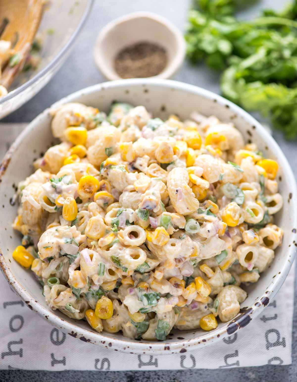 Corn pasta salad with green onions and coriander leaves mixed in mayo dressing served in a white spotted bowl.