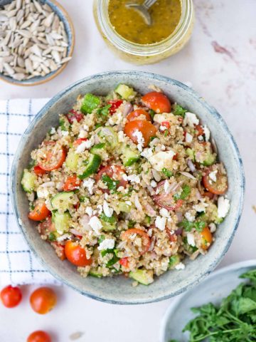 Featured image of Quinoa salad in a bowl along with vinaigrette decoratively put on a table with seeds, herbs and cherry tomato
