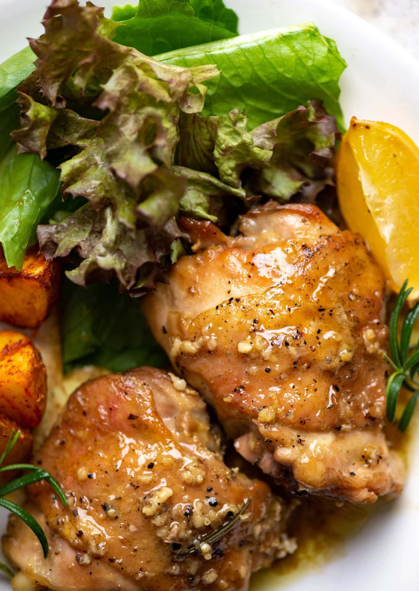 Close up of perfectly baked chicken thighs , done in the oven with a flavorful garlic butter sauce and herby flavor from rosemary. It is served with roasted potatoes and greens.