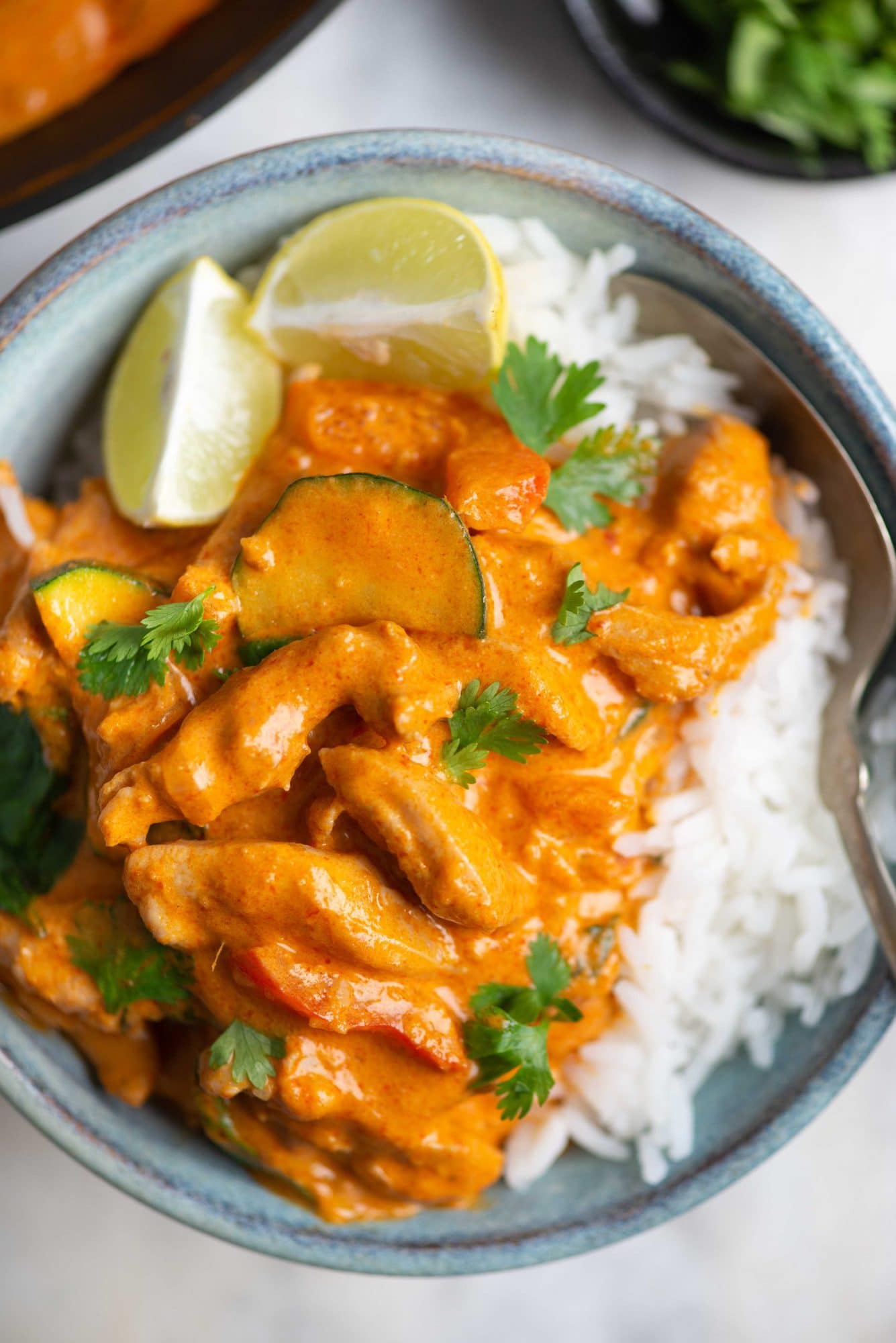 Rich and creamy Thai red Curry served on top of white rice in a blue bowl. Has lemon slice, basil and cilantro as garnish.