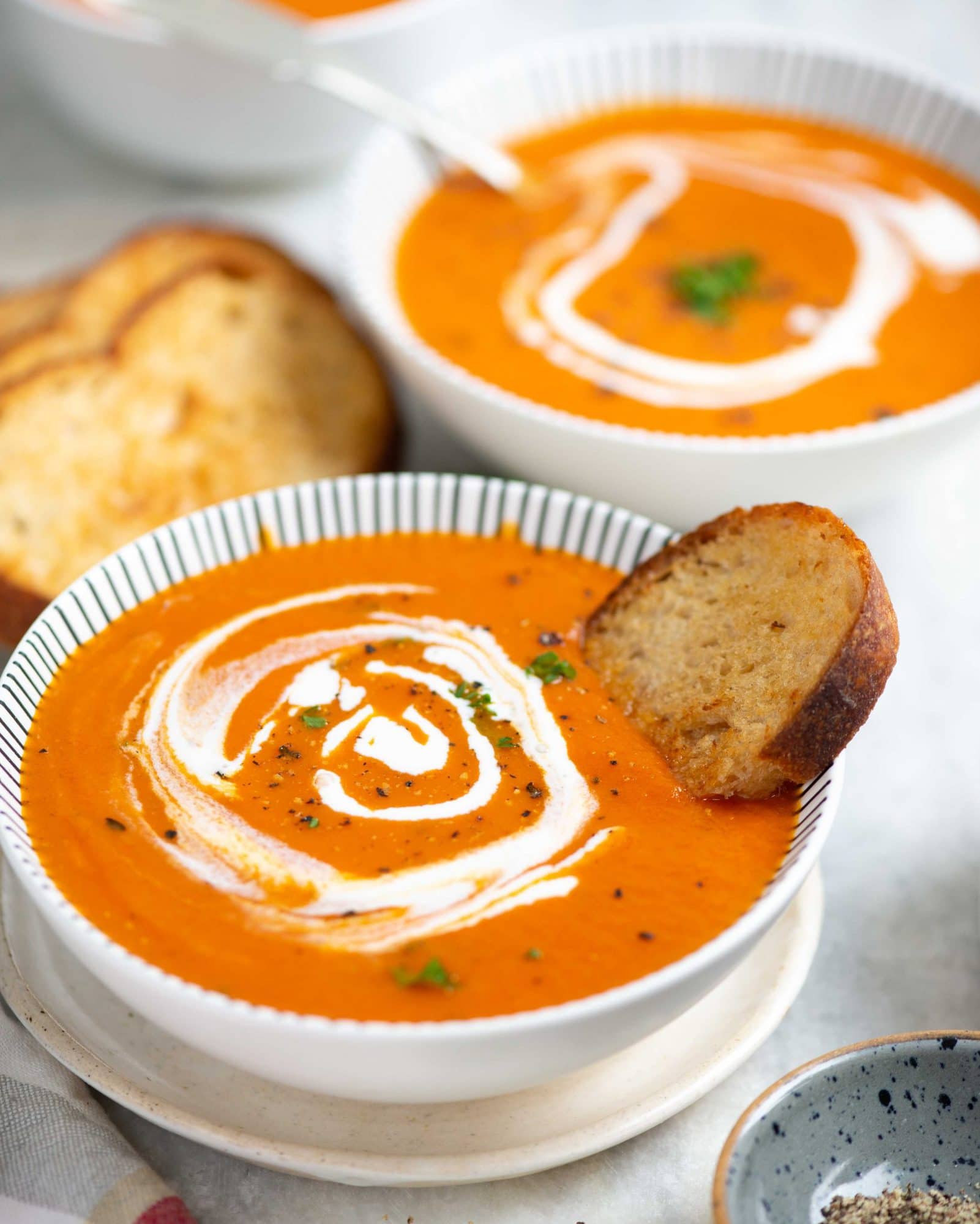 Homemade tomato soup is served in a white bowl with a crouton dipped in it. It is also garnished with basil and cream on top.