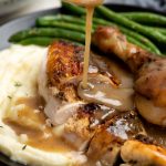 Shot of gravy poured on slices of roast chicken along with mashed potato and green beans on a plate.