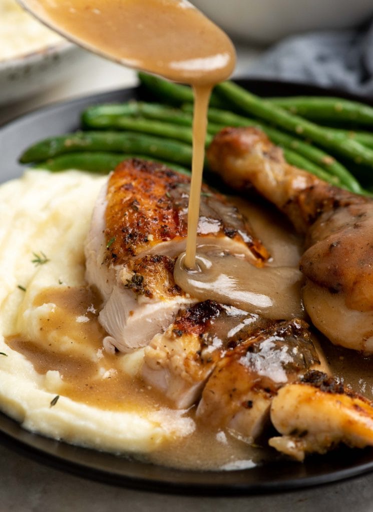 Shot of gravy poured on slices of roast chicken along with mashed potato and green beans on a plate.