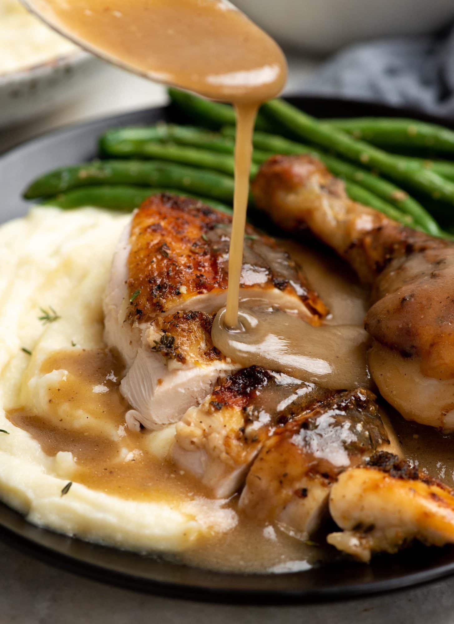 Picture of pouring gravy on a slice of roast chicken with mashed potatoes and green beans on a black plate.