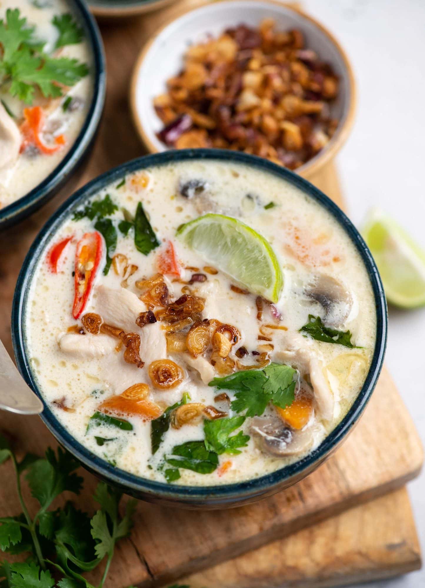 Thai coconut chicken soup or Tom kha gai/kai plated in a blue bowl with chicken, mushrooms, coriander, bird-eye chili shown on top and garnished with fried garlic and lime slice.