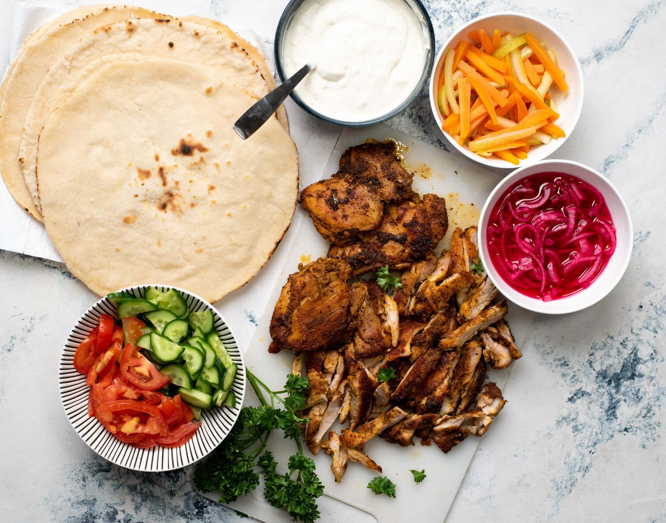 Shows what goes into chicken shawarma - Pita bread, grilled chicken, garlic sauce, pickled carrots and onions, chopped tomatoes and cucumber.