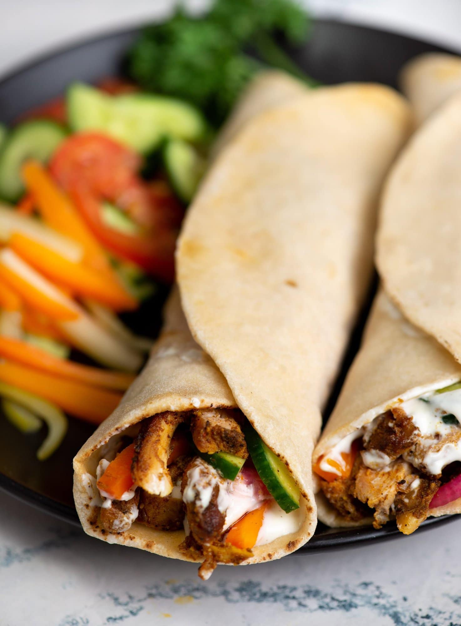 Cover pic of chicken shawarma showing couple of shawarma wrap beside chopped veggies on a black plate and the stuffing looks creamy.