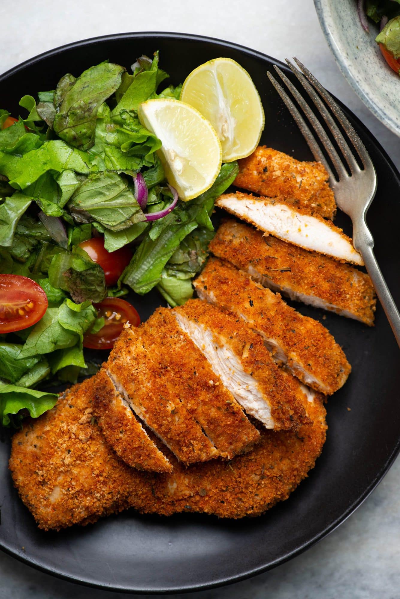 Sliced crispy crusty chicken breasts served with chopped lettuce, cherry tomatoes and lemon on a black plate.