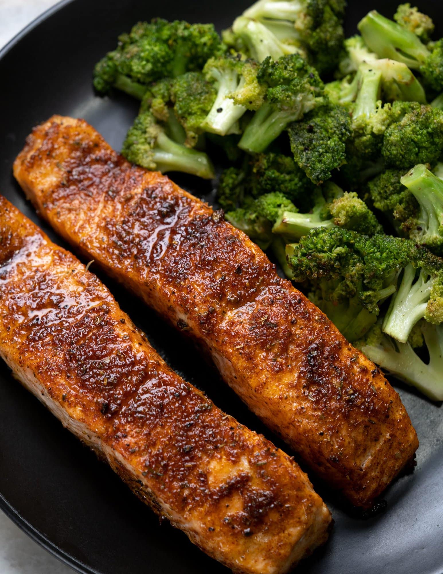 Air fried salmon with a crusty skin on top, served with roasted broccoli on a black plate.