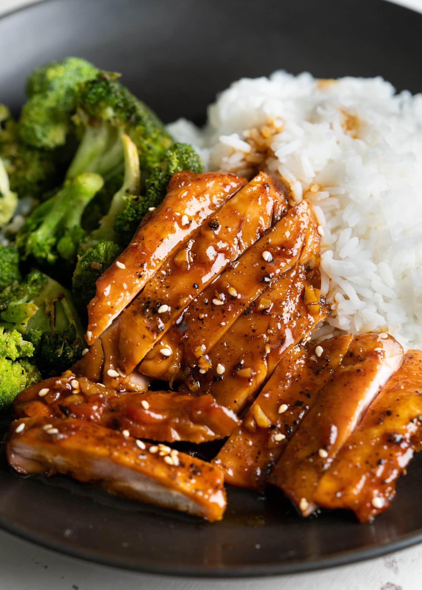 Sliced chicken coated with honey garlic sauce and served with rice and broccoli on a black plate.