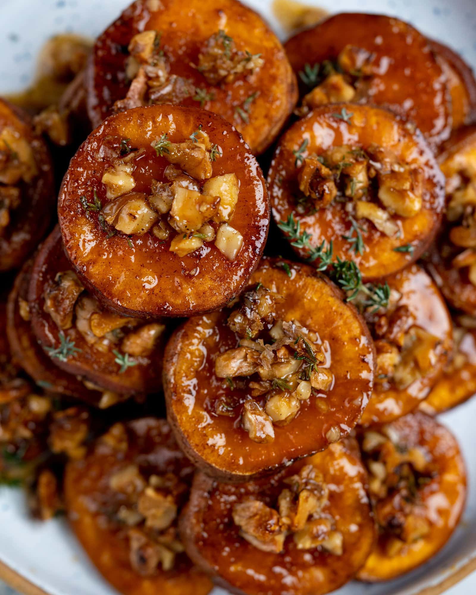 Bunch of roasted sweet potatoes stacked as a heap. These candied sweet potatoes have a glaze from the maple syrup and have crunchy walnuts on top with a twig of thyme on the bunch.
