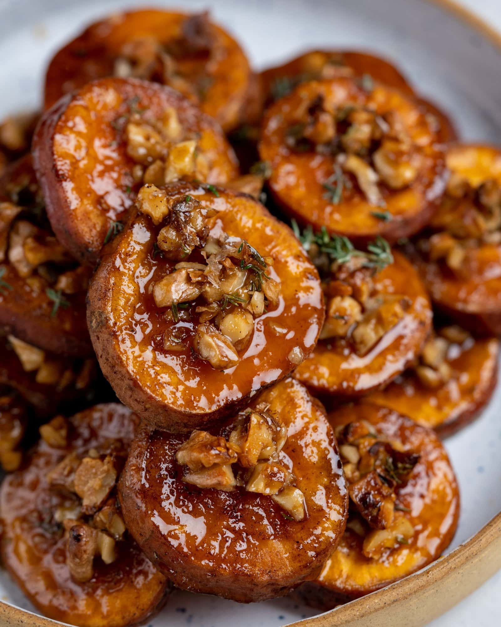 A heap of roasted sweet potatoes served on a white plate. Close up shows syrupy glaze on the potatoes with crunchy walnuts, taste of cinnamon and flavor of thyme on them.