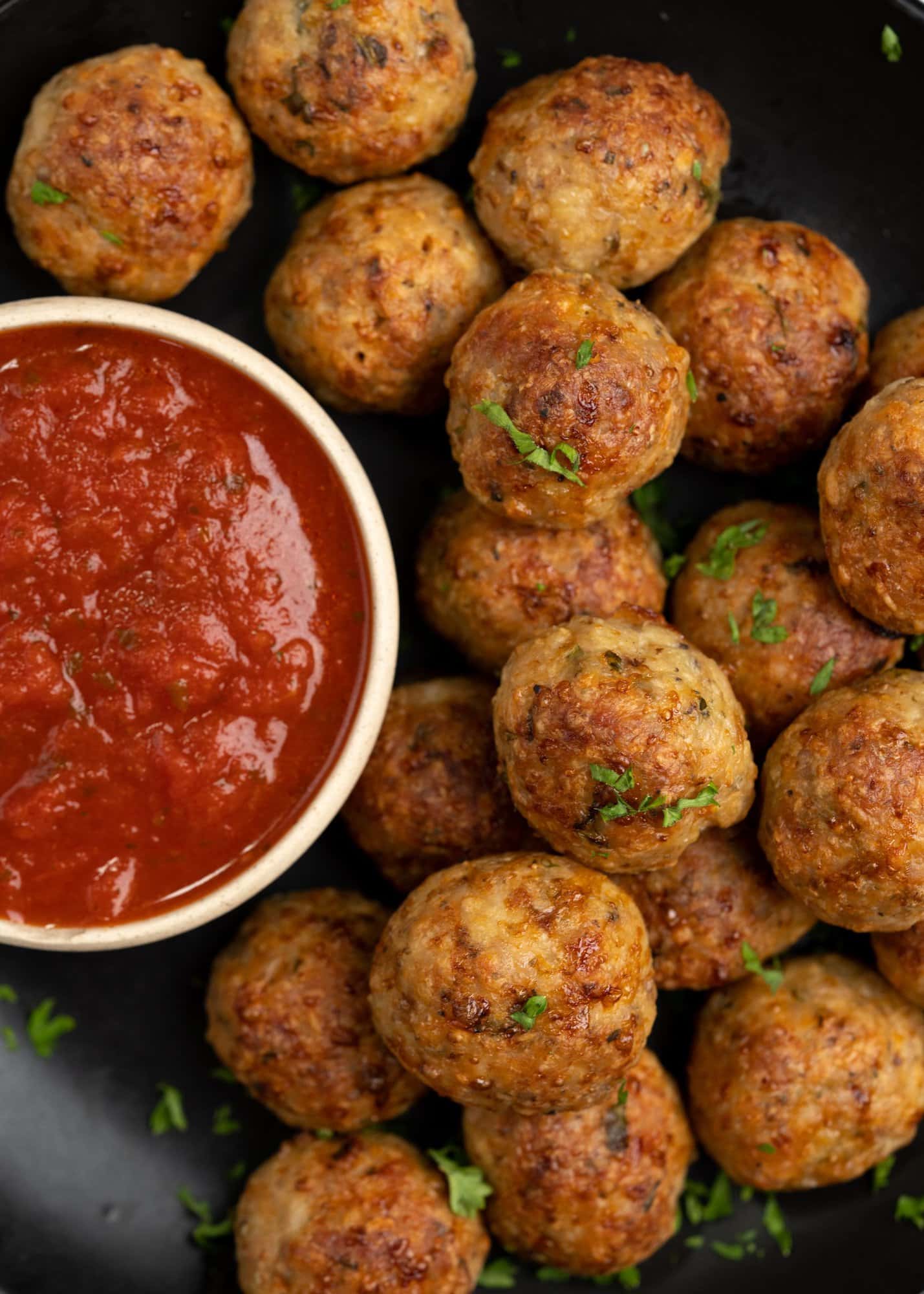 Golden brown chicken meatballs, with a crisp exterior and juicy chicken inside, are baked and served on a black plate, along with marinara sauce.