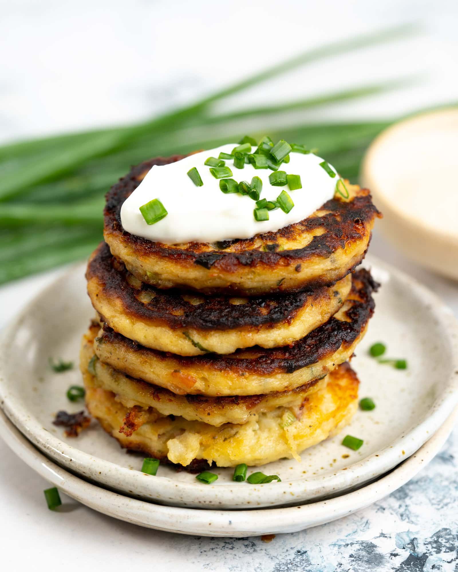 Mashed Potato Pancakes stacked on plates with sour cream and chopped green onions on top.