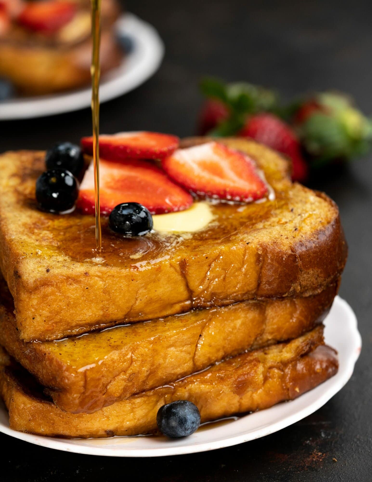 Maple syrup being drizzled over a stack of french toasts with toppings of berries and butter.