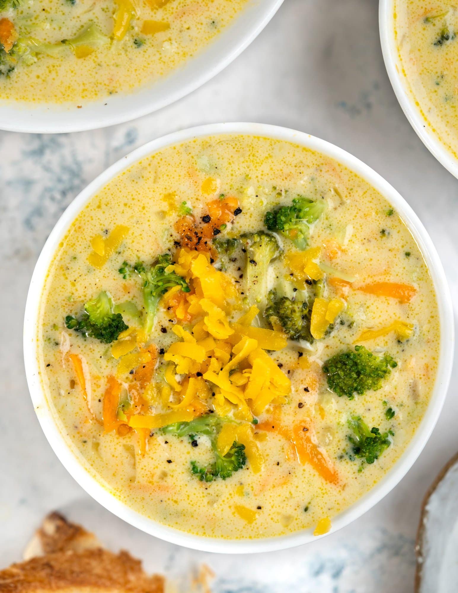 Shredded cheddar and pepper is sprinkled on top of broccoli cheddar cheese soup, served inn a white soup bowl.