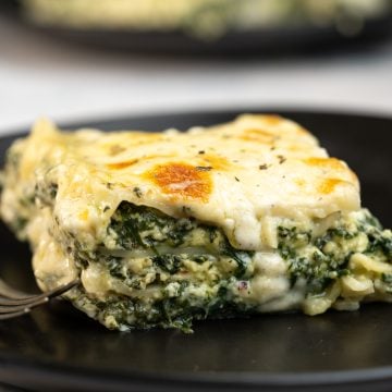 A slice of spinach lasagna with cheese oozing out of the layers and served on a black dining plate.