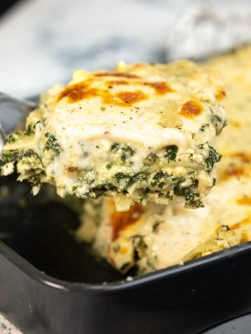 A slice of classic spinach lasagna picked up with a slotted spoon. The lasagna is creamy and cheesy between the layers with a white sauce.