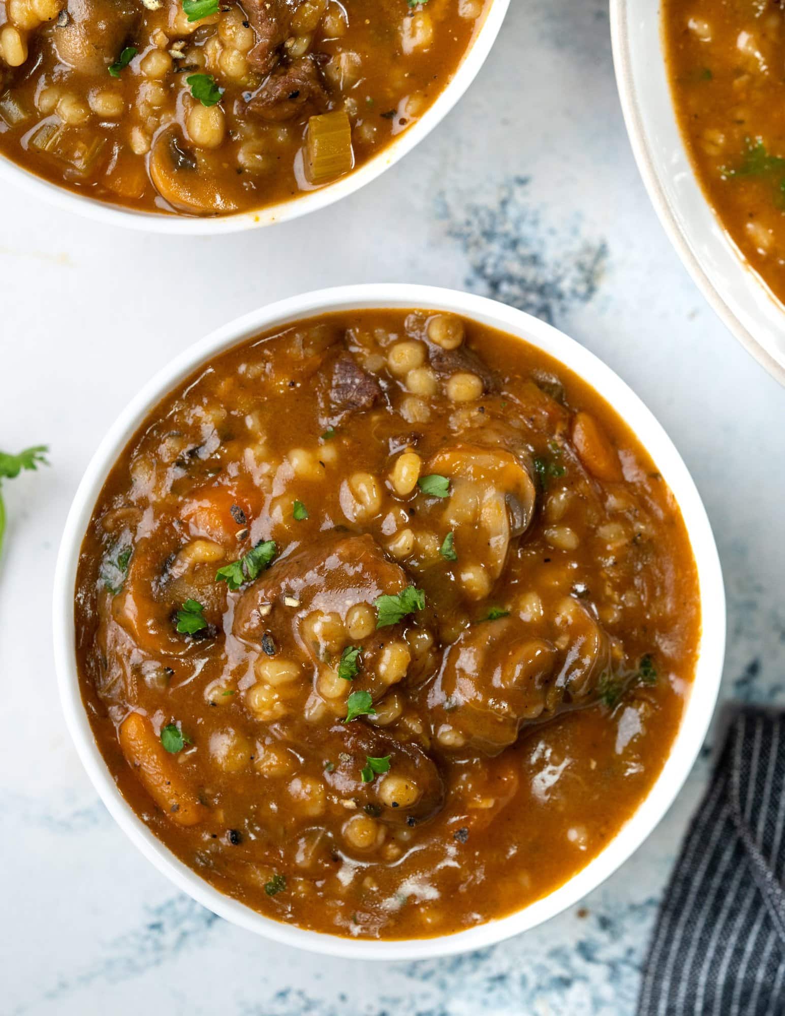 Beef barley soup served in a white bowl with chopped fresh parsley sprinkled on top.