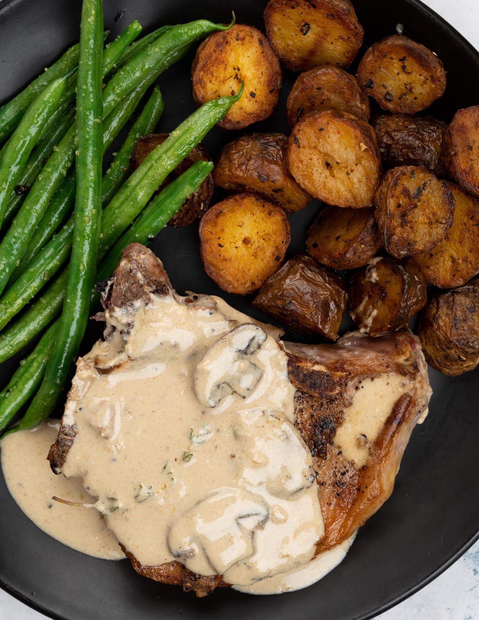 Creamy mushroom sauce drizzled over a pork chop with brown crust and served on a black plate with roasted potatoes and sauteed green beans.