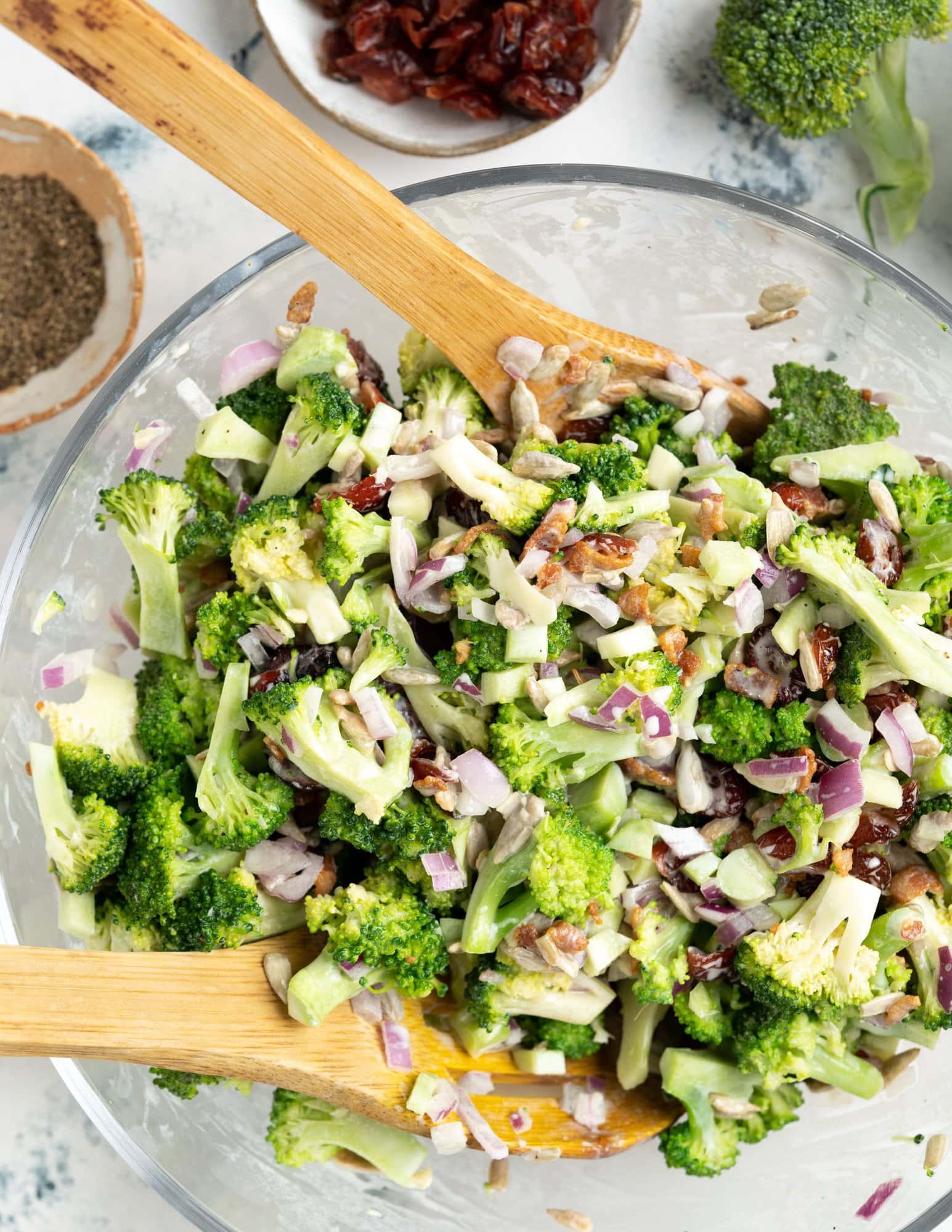 Fresh crisp broccoli, bits of onion and bacon, seeds and cranberry tossed in a creamy yogurt-based dressing, in a large mixing bowl with a wooden spatula.