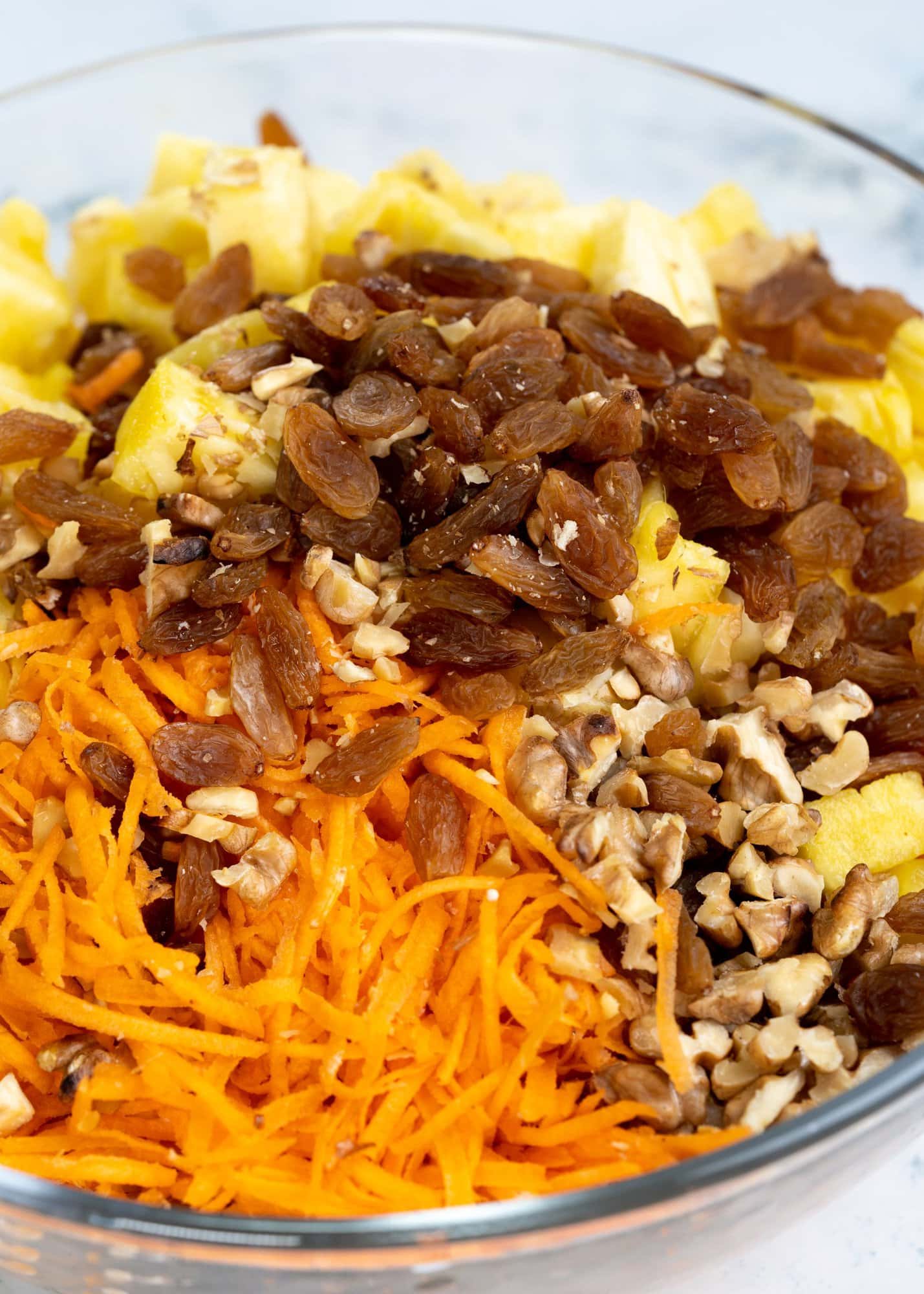 Shows all the ingredients to make carrot salad - shredded carrots, raisins, chopped pineapple and toasted walnuts pieces in a bowl.