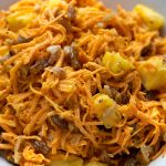 Close up view shows shredded carrots, raisins, chopped walnuts and pineapple chunks coated with creamy mayo-based dressing having dijon mustard.