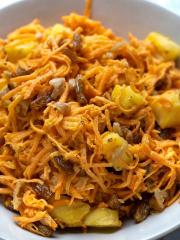 Close up view shows shredded carrots, raisins, chopped walnuts and pineapple chunks coated with creamy mayo-based dressing having dijon mustard.