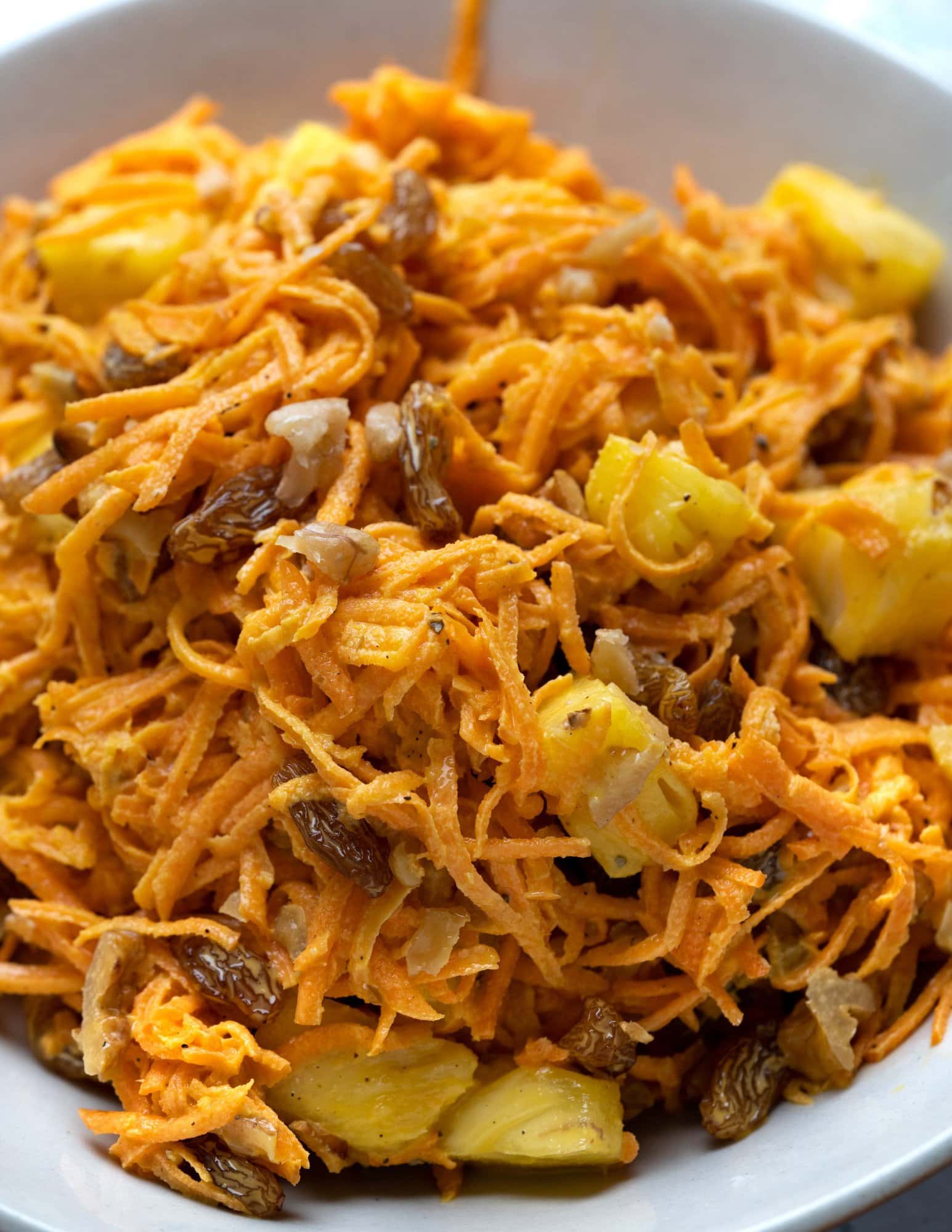 Close view of creamy carrot salad shows shredded carrots, raisins, chopped pineapple and walnuts coated in a creamy mayo dressing with mediterranean flavors.