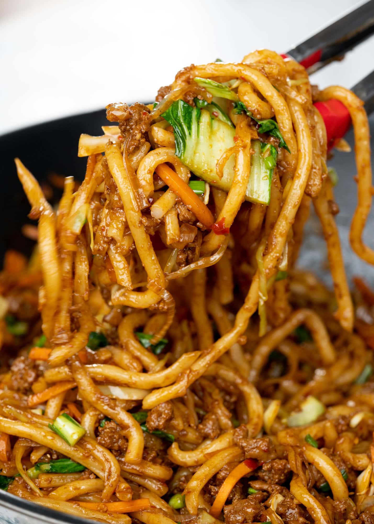 Close view of stir-fried udon noodles picked with a tong. Pickings has noodles coated with sauce, ground pork and pieces of bok choy.