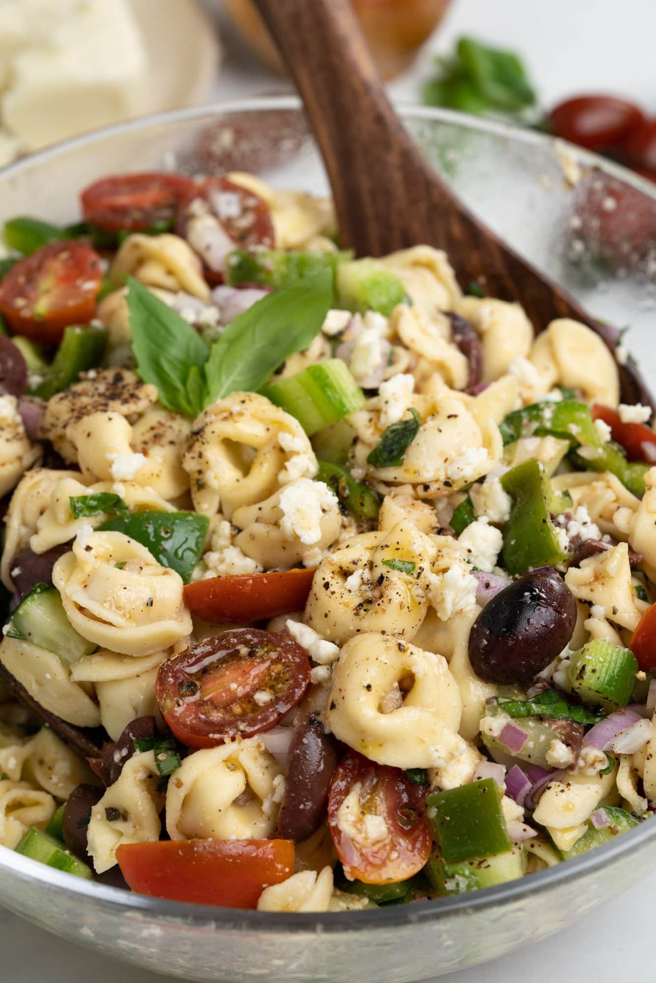 Close up of Greek Tortellini Pasta salad, being tossed with a wooden spoon in a glass bowl.