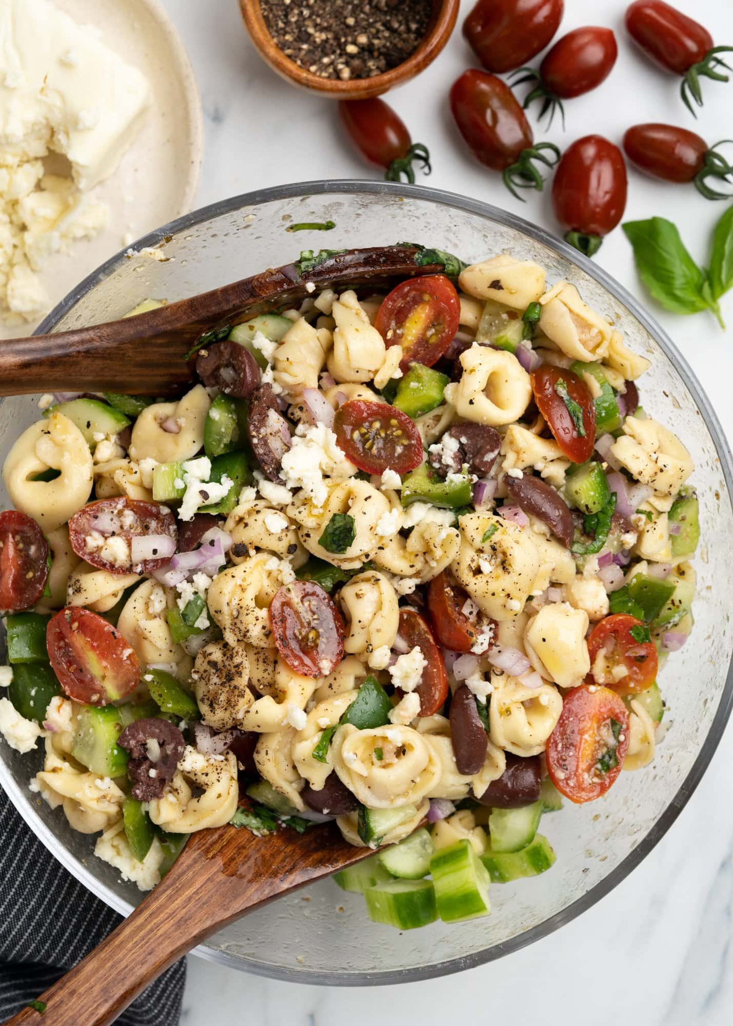 Tortellini pasta salad with greek dressing and feta sprinkled on top in a glass bowl with a wooden spatula.