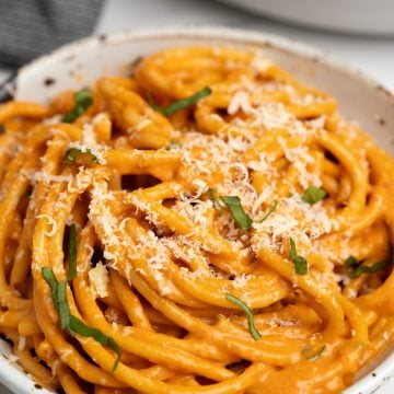 Image shows red pepper pasta served in a white bowl with shredded parmesan and basil on top.