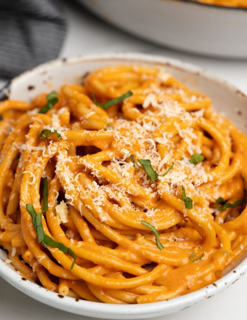 Image shows red pepper pasta served in a white bowl with shredded parmesan and basil on top.