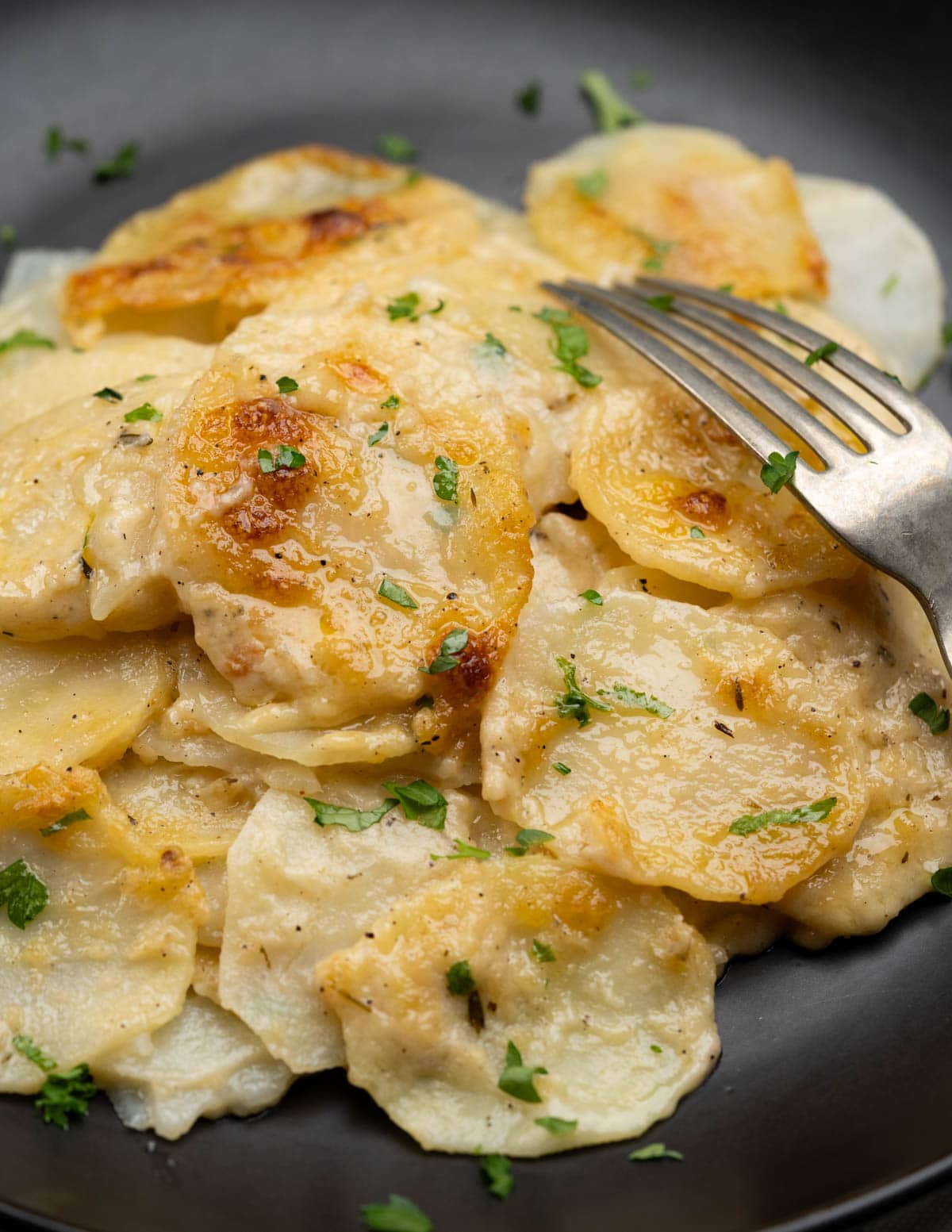 Tender potato slices coated with a creamy sauce served on a black plate and fork and topped with a few slices that have developed a nice crust.