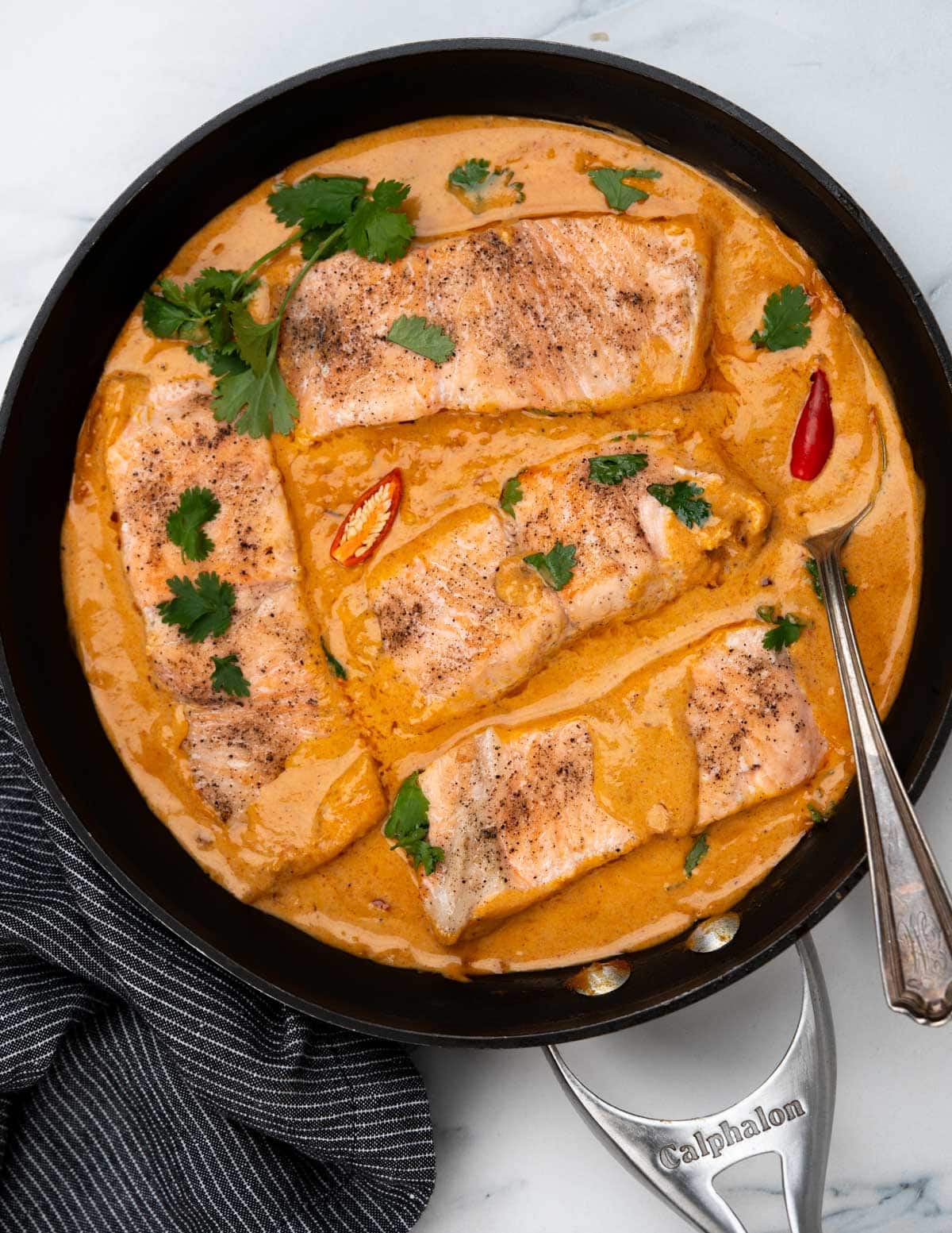 Coconut salmon curry made in a black skillet, shows four salmon fillets engulfed in a creamy rich coconut cury sauce and garnished with cilantro and chili peppers.