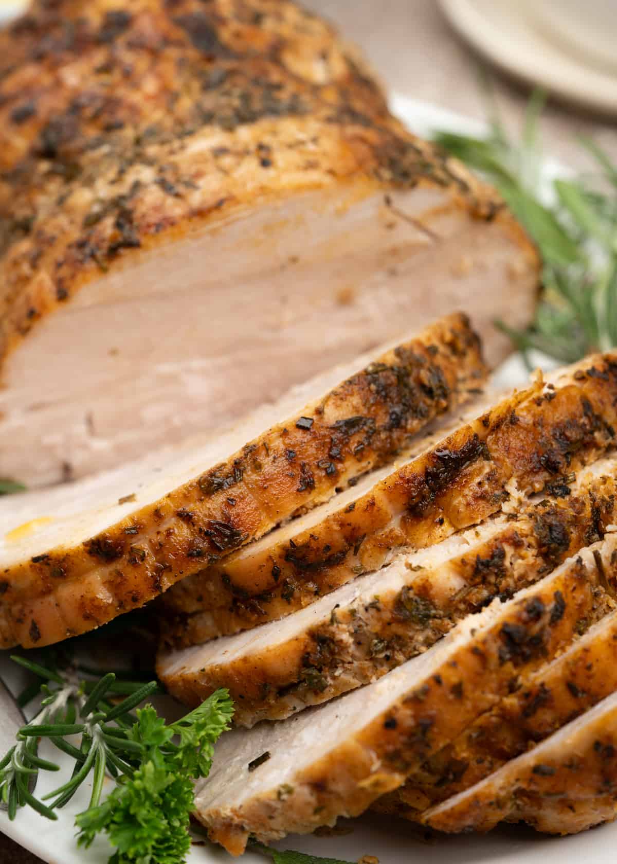 Shows a stack of sliced turkey breast roast highlighted a herby crust and tender meat and garnishes with rosemary sprig.
