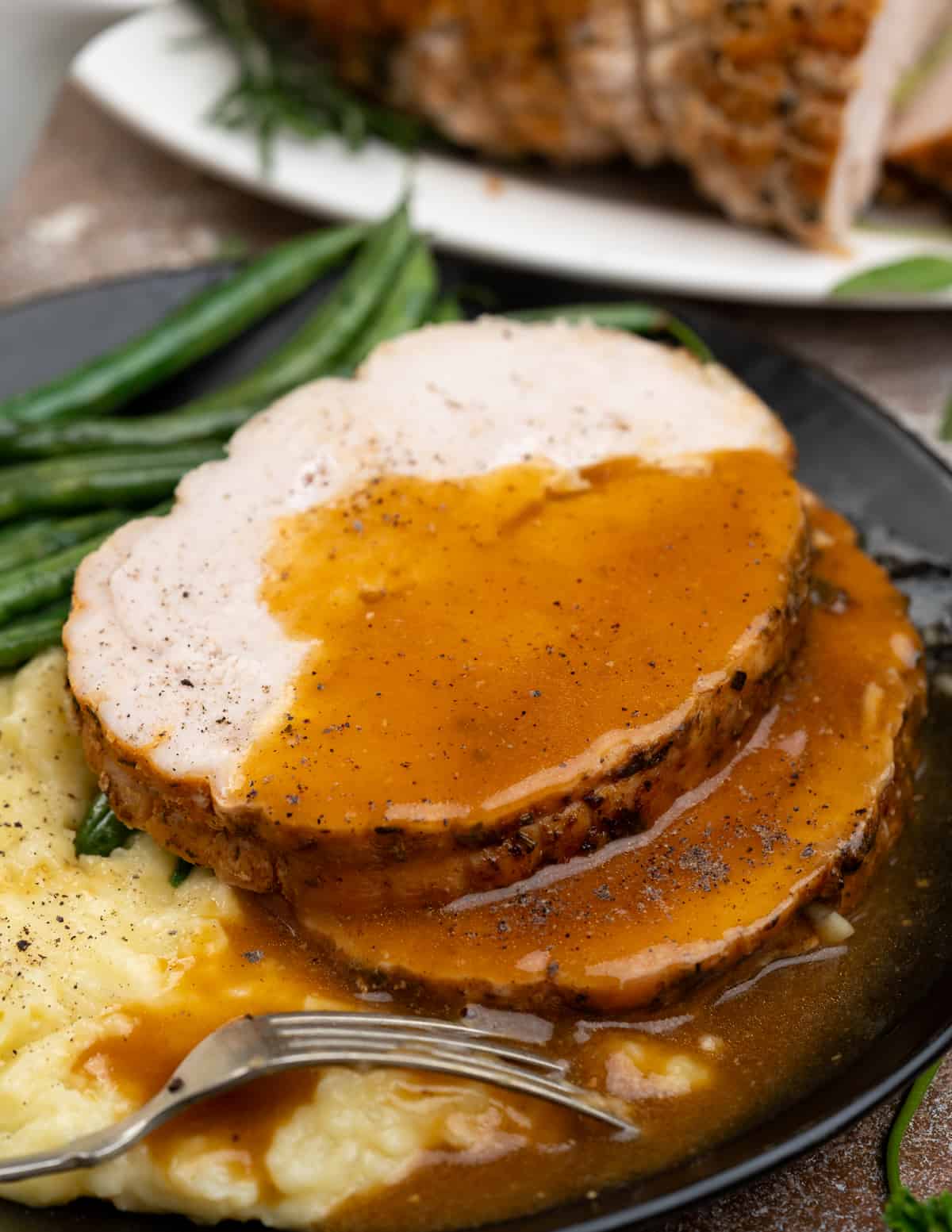 Two slices of roasted turkey breast served with a flavorful, velvety gravy along with mashed potatoes and green beans on a black plate.