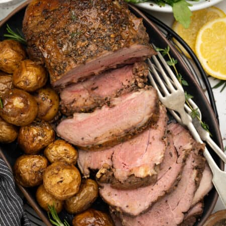 Lamb roast sliced and plated along with roasted potatoes, herbs and forks.