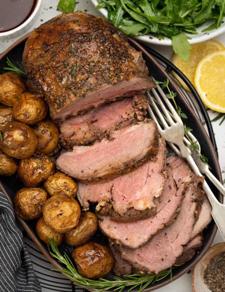 Lamb roast sliced and plated along with roasted potatoes, herbs and forks.