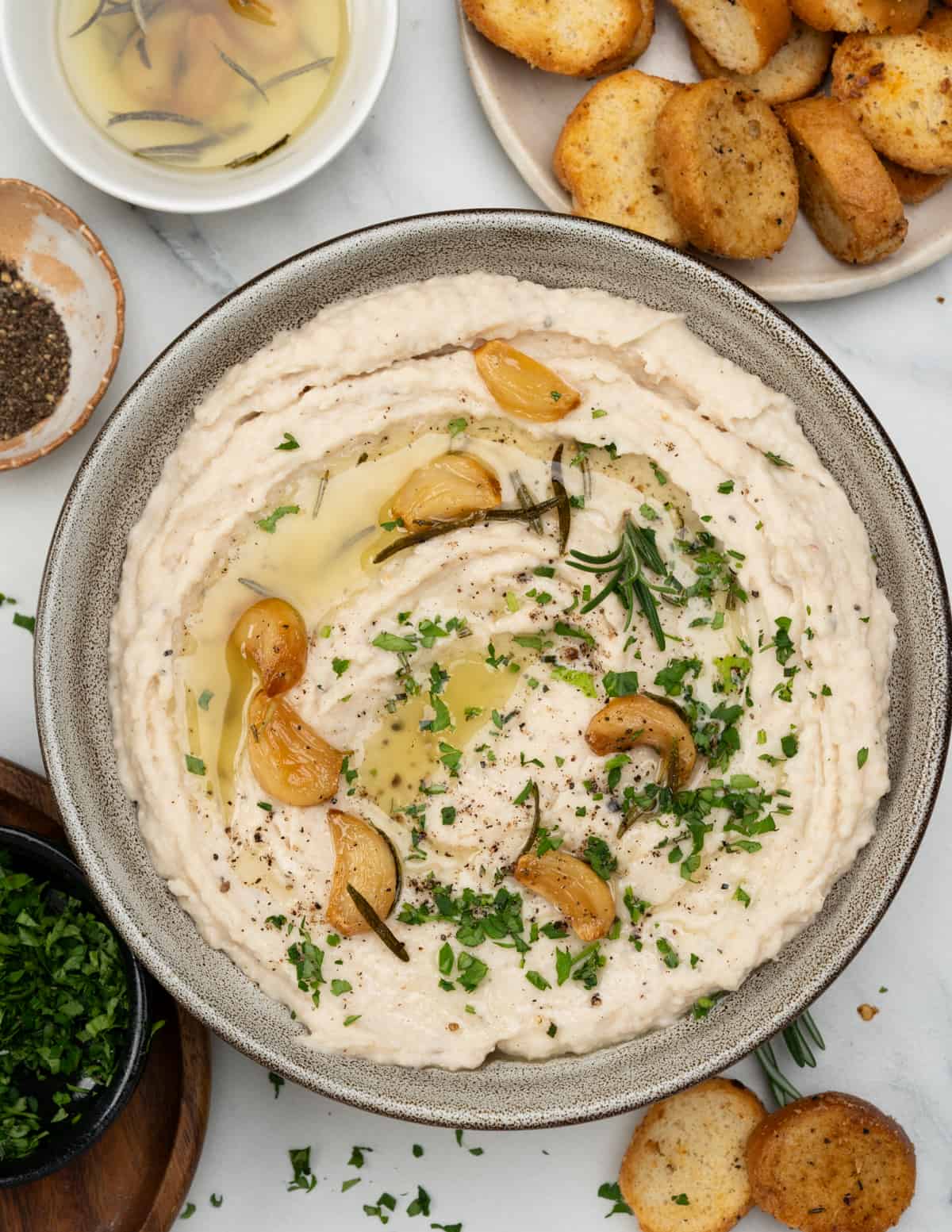 White bean dip drizzled with garlic and rosemary oil, topped with roasted garlic cloves and garnished with chopped parsley.