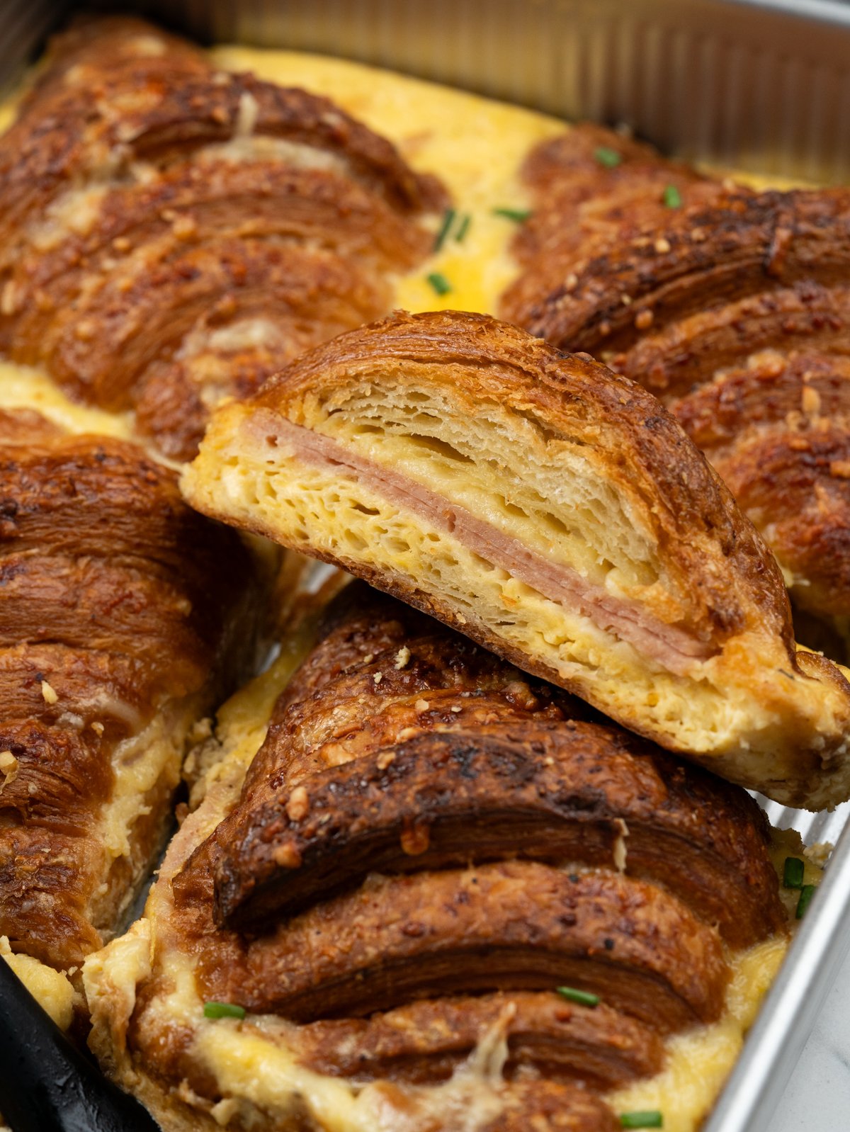 Cross section of croissant breakfast casserole showing flaky croissant, ham, melted cheese and egg. milk layer.